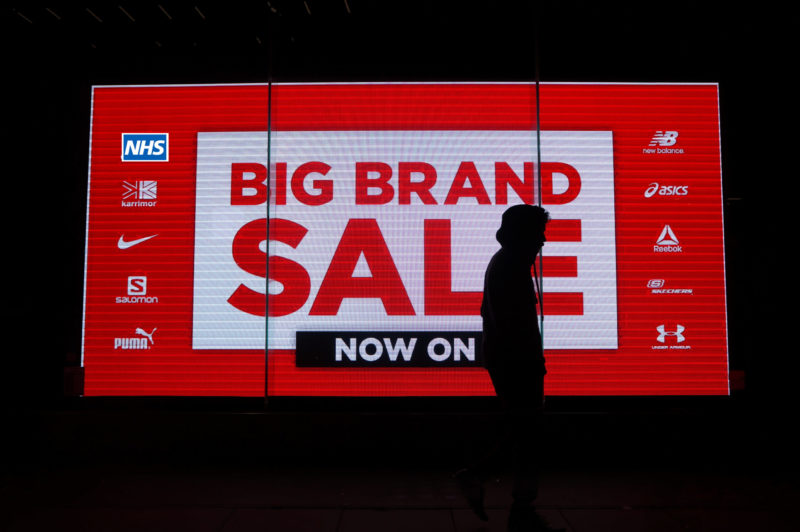 Image of illuminated billboard at night saying "Big Brand Sale" and showing various major brand logos including the NHS