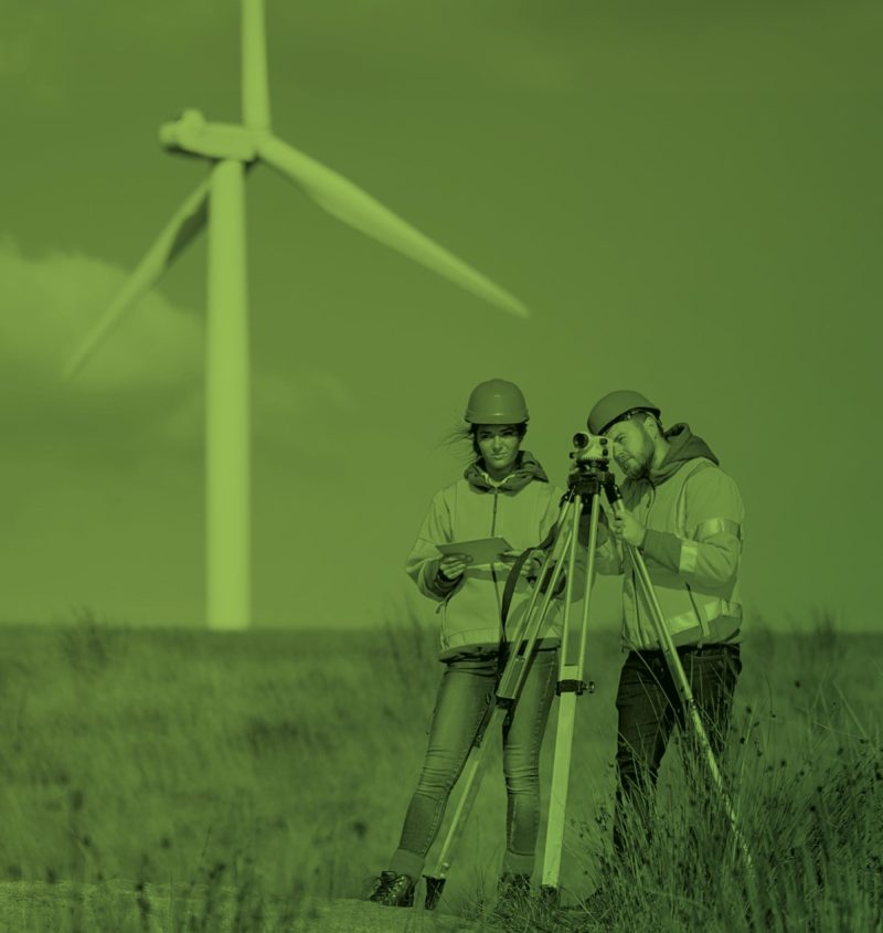 Two land surveyors in a large expanse of countryside with a wind turbine in the background 